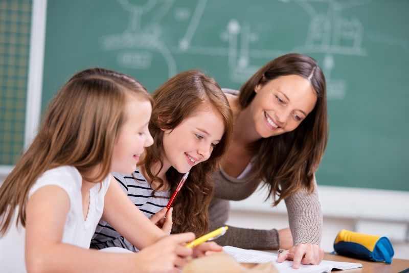 A teacher is helping two young girls with their homework in a classroom.