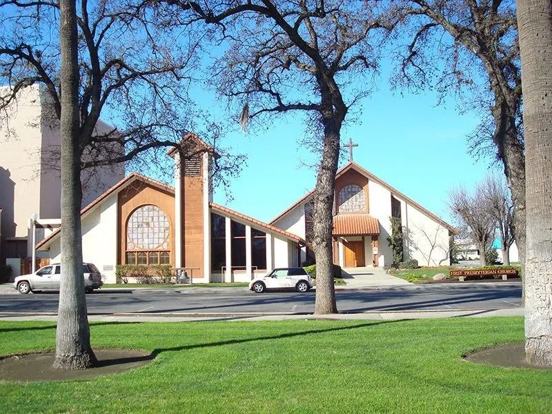 Church building with bell tower, trees, green lawn, and parked cars on a sunny day.