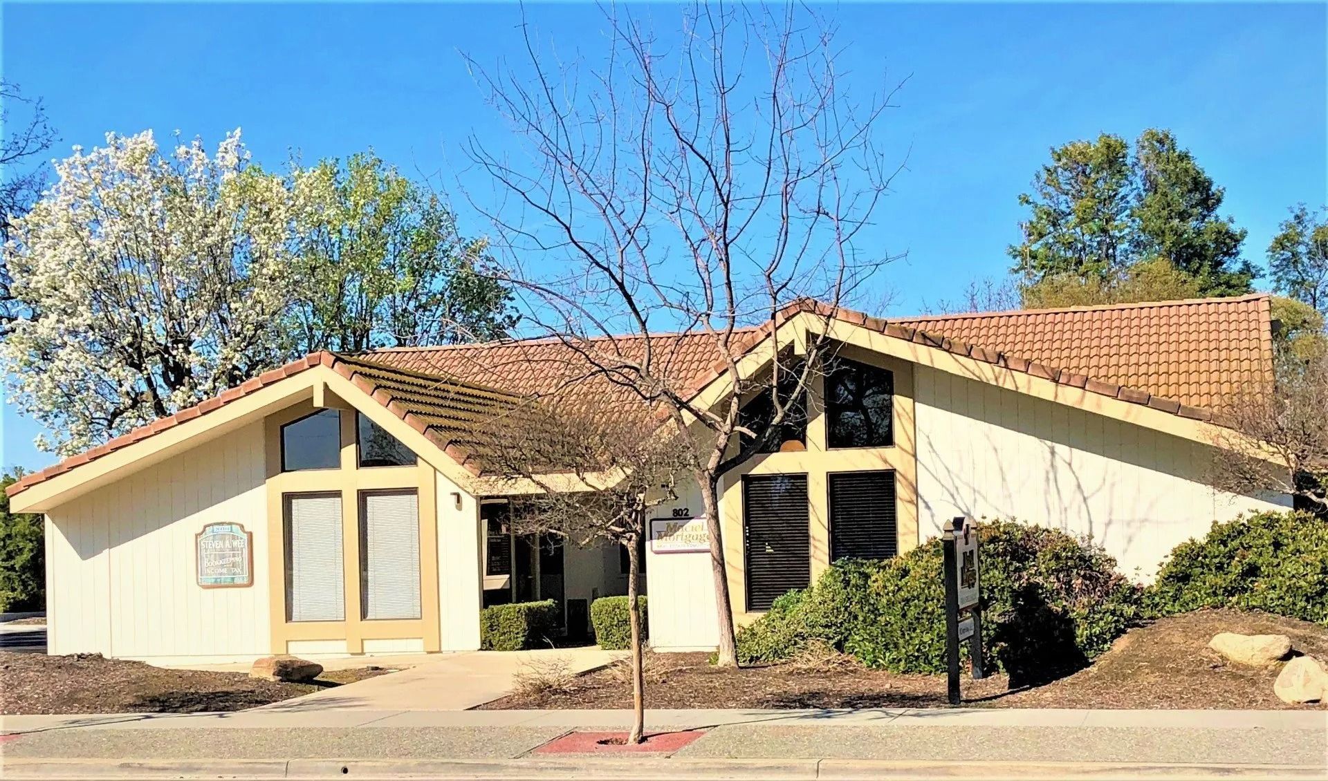 Tan building with brown tiled roof, large windows, and surrounding greenery under a blue sky.