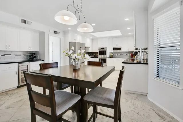 Bright white kitchen with dark wood dining table and chairs, stainless steel appliances, and marble flooring.