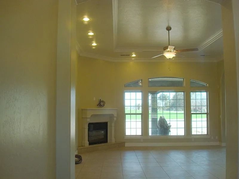Empty living room with fireplace and large windows, yellow walls, tile floor, and ceiling fan.
