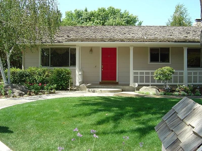 House with red door, covered porch, and green lawn. Wooden shake roof.