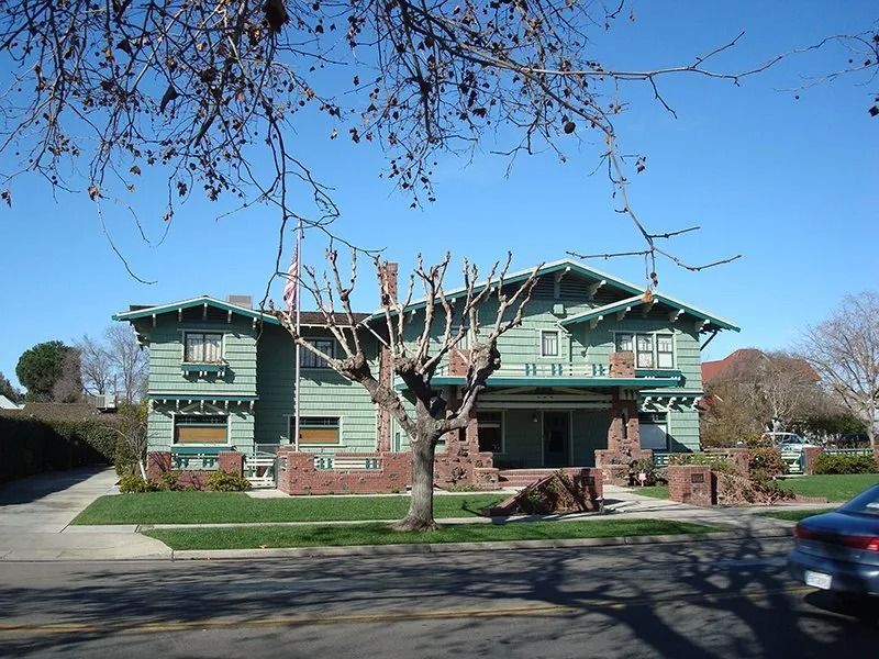 Two-story green Craftsman house with brick accents, blue sky, and a car parked on the street.