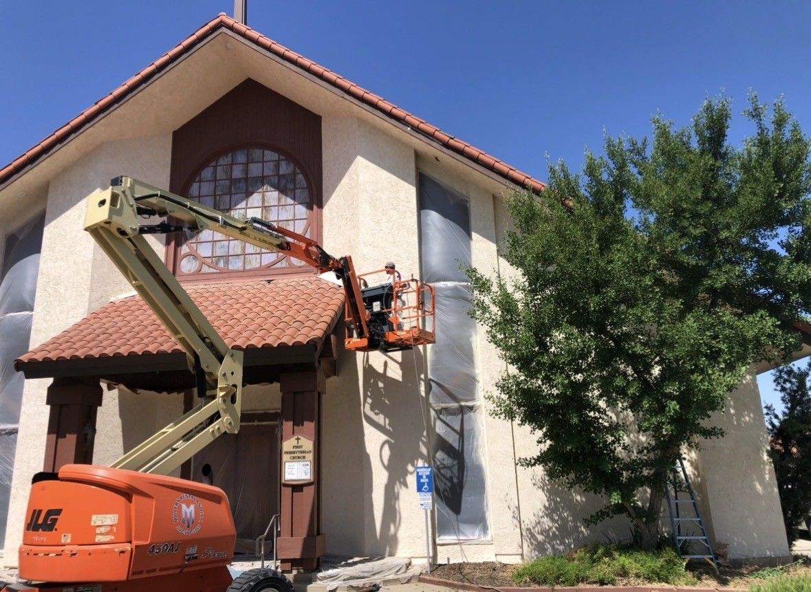 Worker in a lift painting a building's exterior; orange lift, beige building, blue sky.