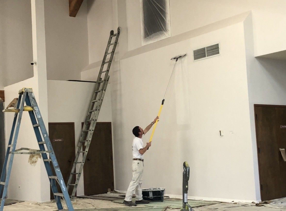 Man painting a high white wall with a roller, using a long extension pole. Ladders nearby.