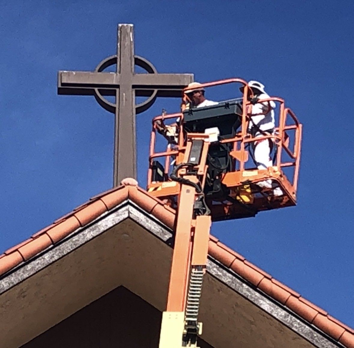 Two workers in a lift platform near a church cross, against a blue sky.