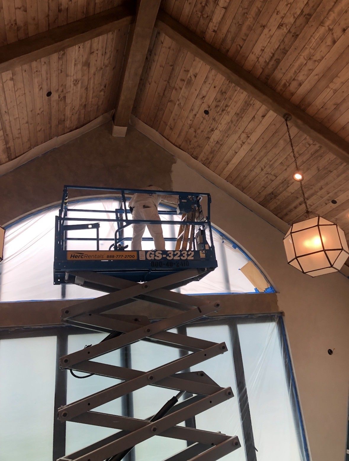 Person painting an arched wall from a scissor lift inside a room with a wood-beam ceiling.