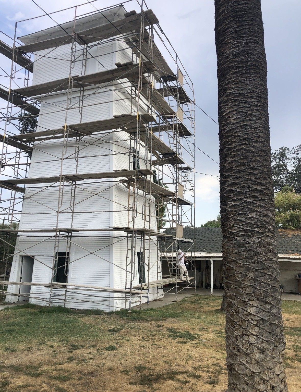White tower under renovation, surrounded by scaffolding, next to a building and palm tree.