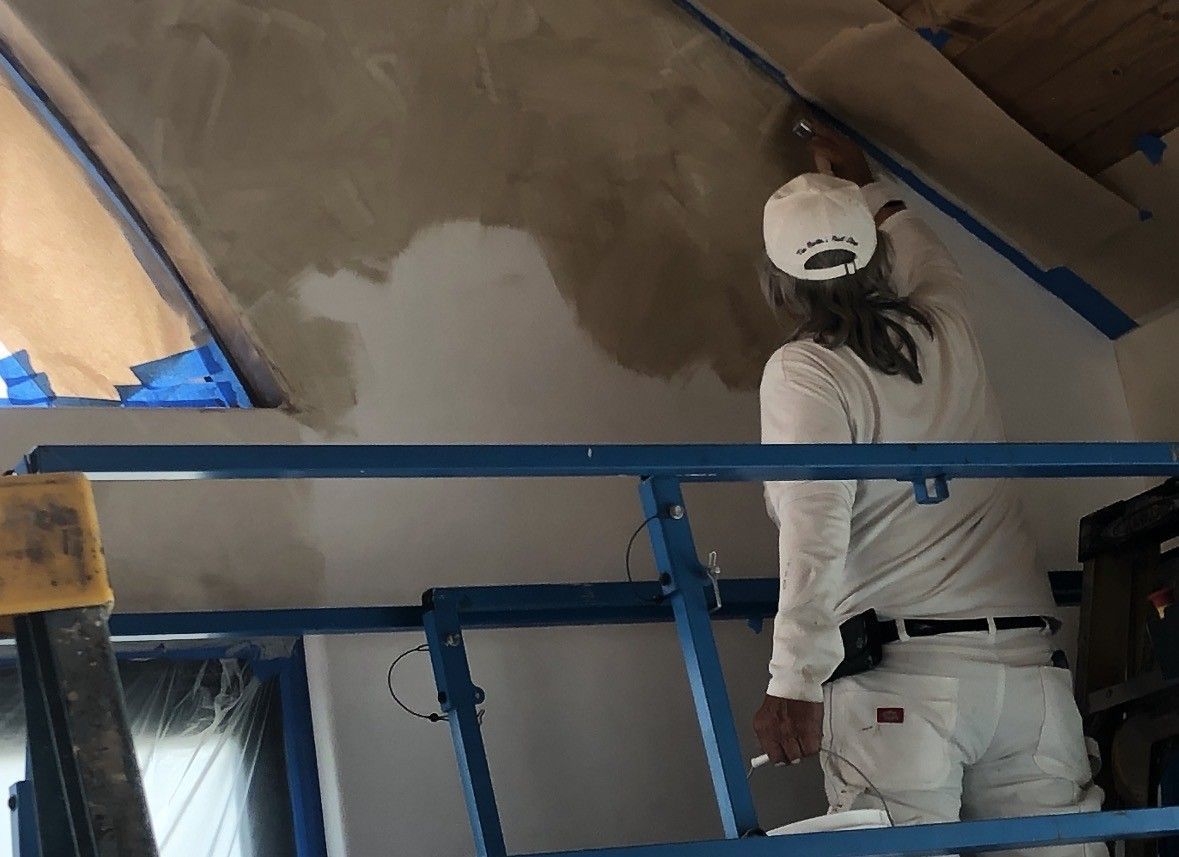 Person painting a wall, standing on a blue lift. Wall is partially painted beige; window with blue tape is visible.