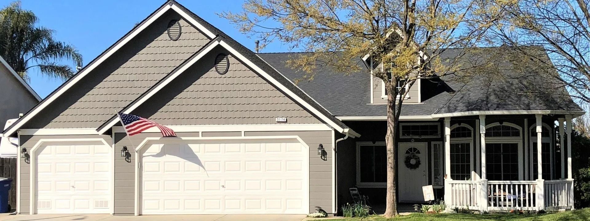 A house with a gray exterior, two-car garage, and a white front porch with a small American flag hanging.