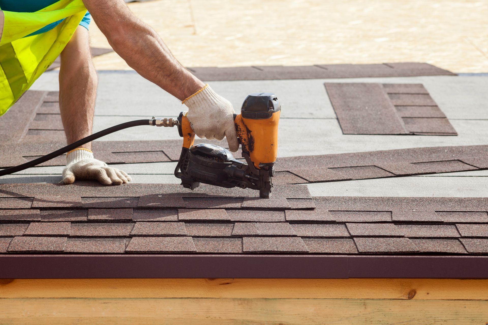 Roofer using a nail gun to install brown asphalt shingles on a roof.