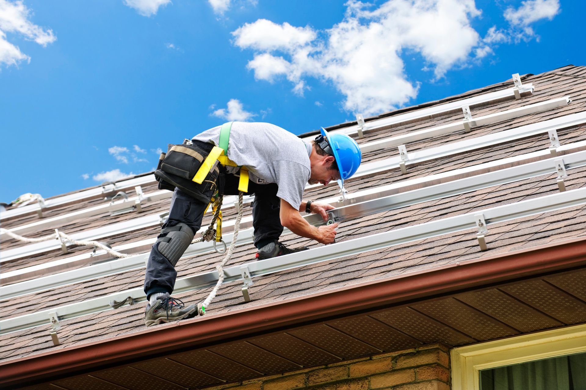 Rooftop solar panel installer, wearing safety harness, secures rails on a brown shingled roof under a blue sky.