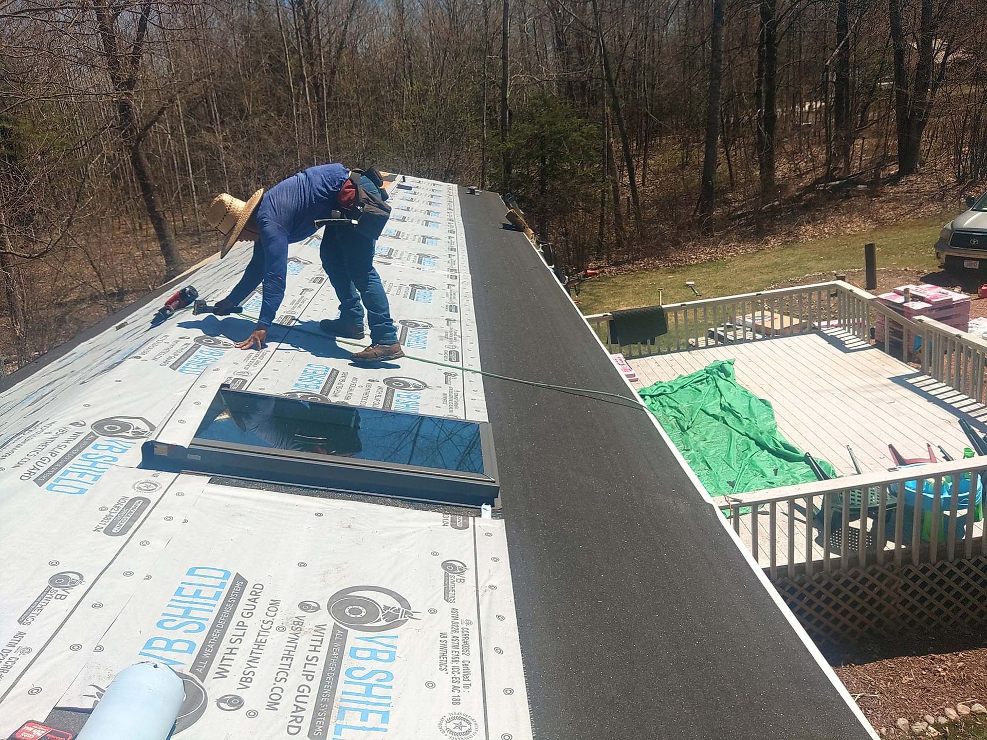 Two workers on a rooftop installing shingles on a sunny day. A deck is visible nearby.