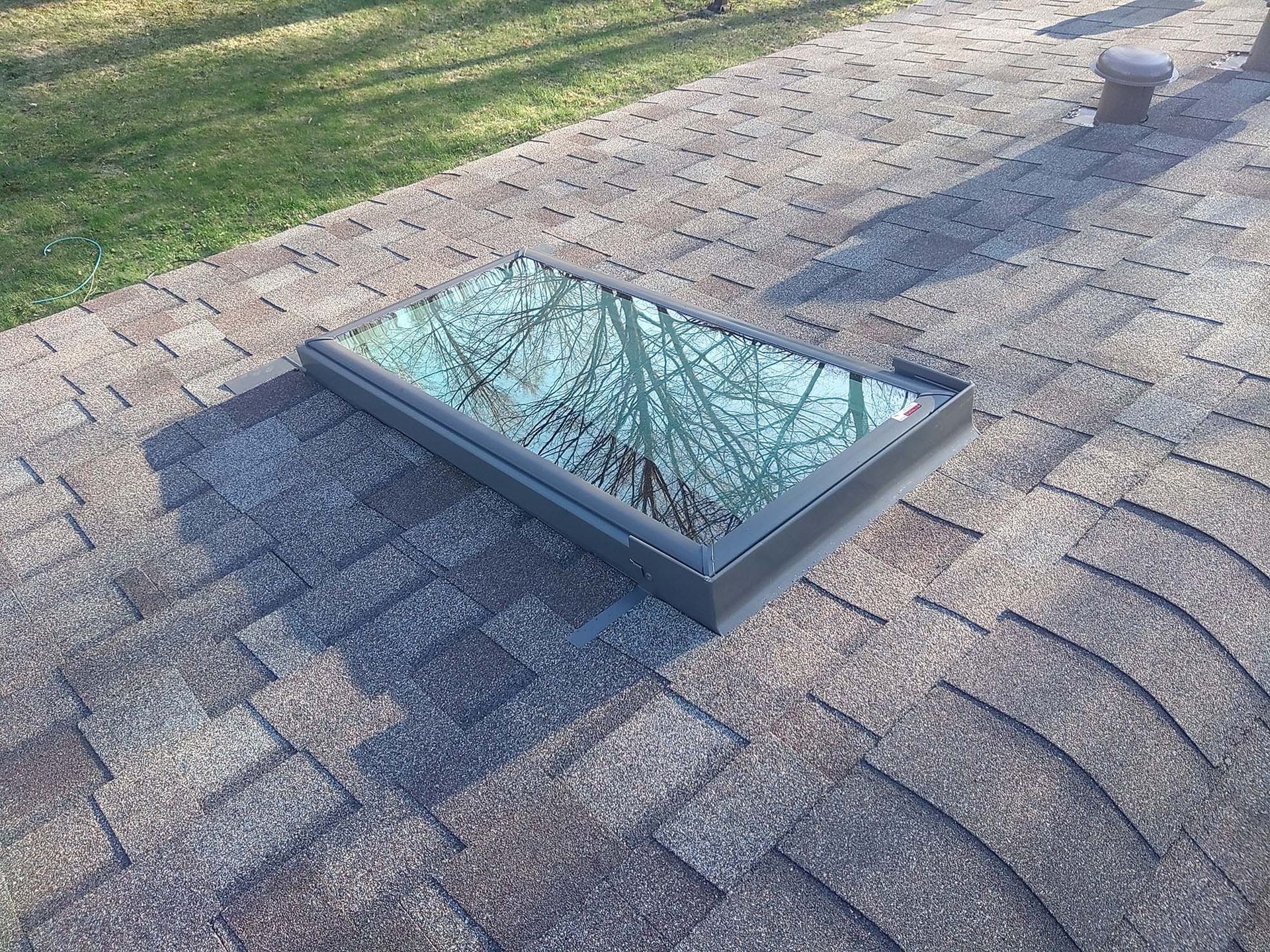 Rectangular skylight on a shingled roof, reflecting trees and sky.