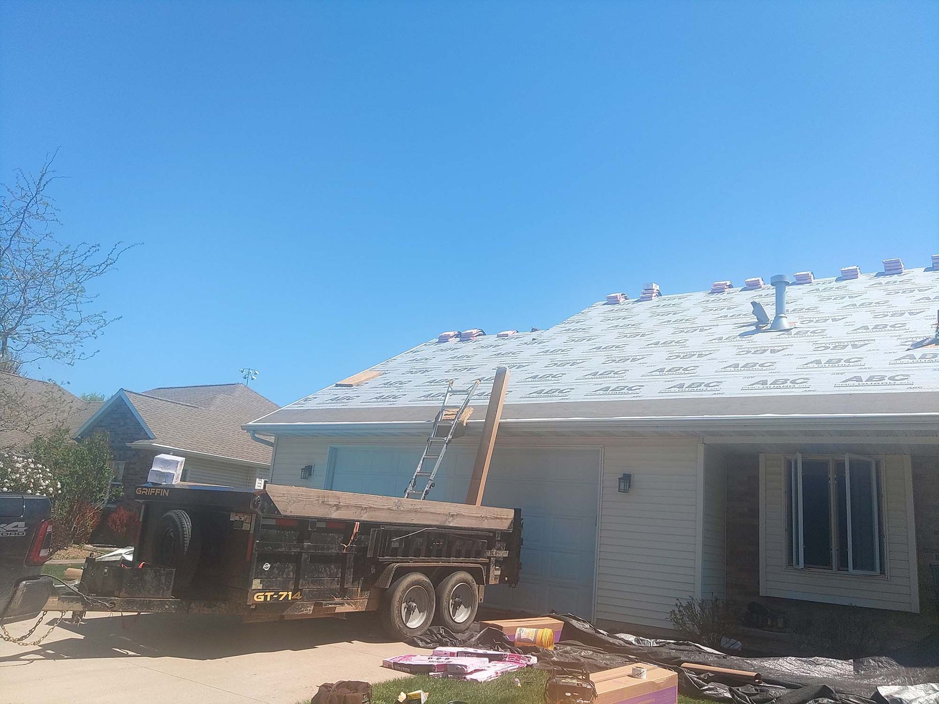 Roofing work on a house; trailer parked nearby. Blue sky, shingles removed.