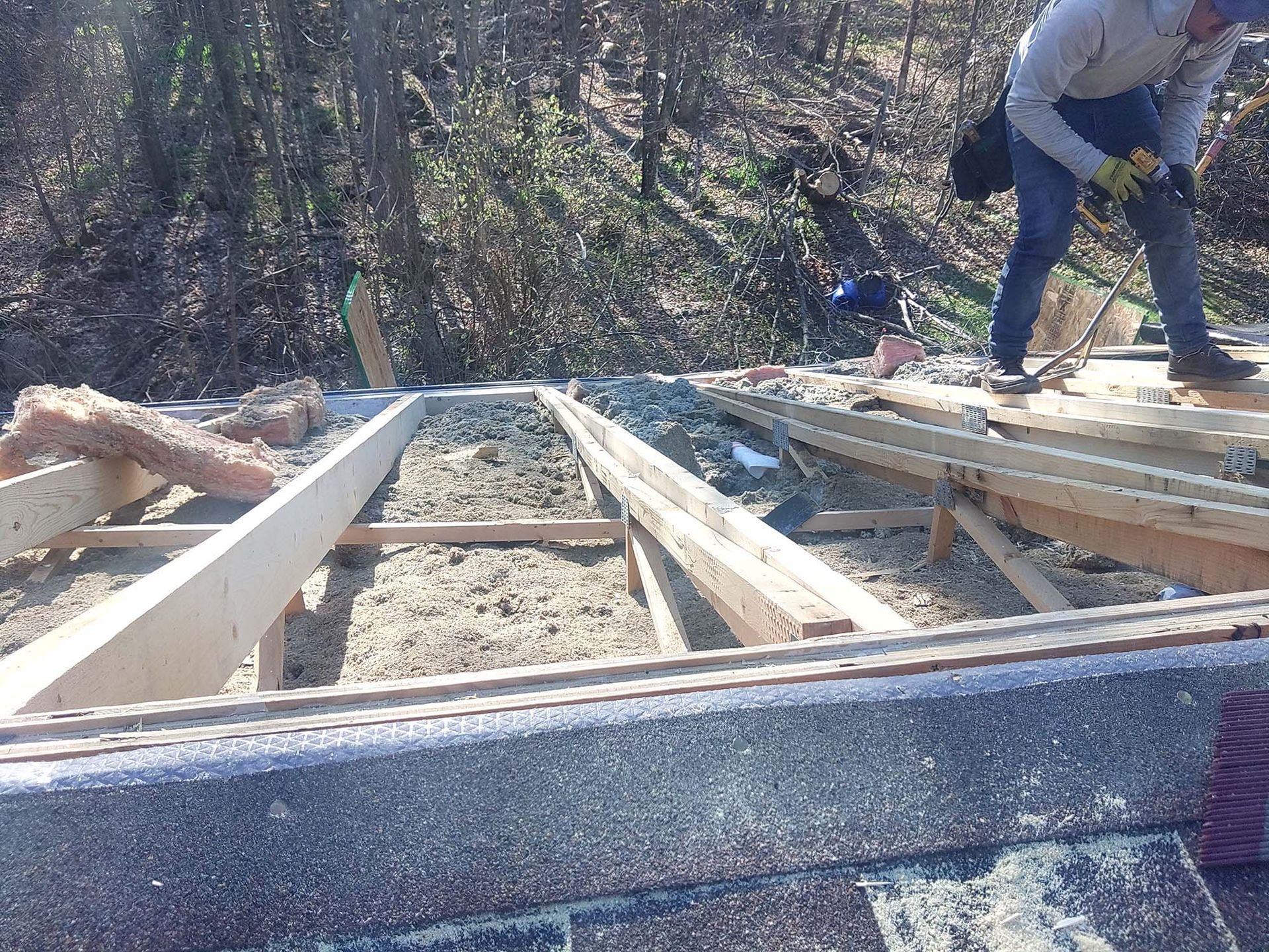 Man on roof using a tool; wood frame with insulation visible; trees in background.