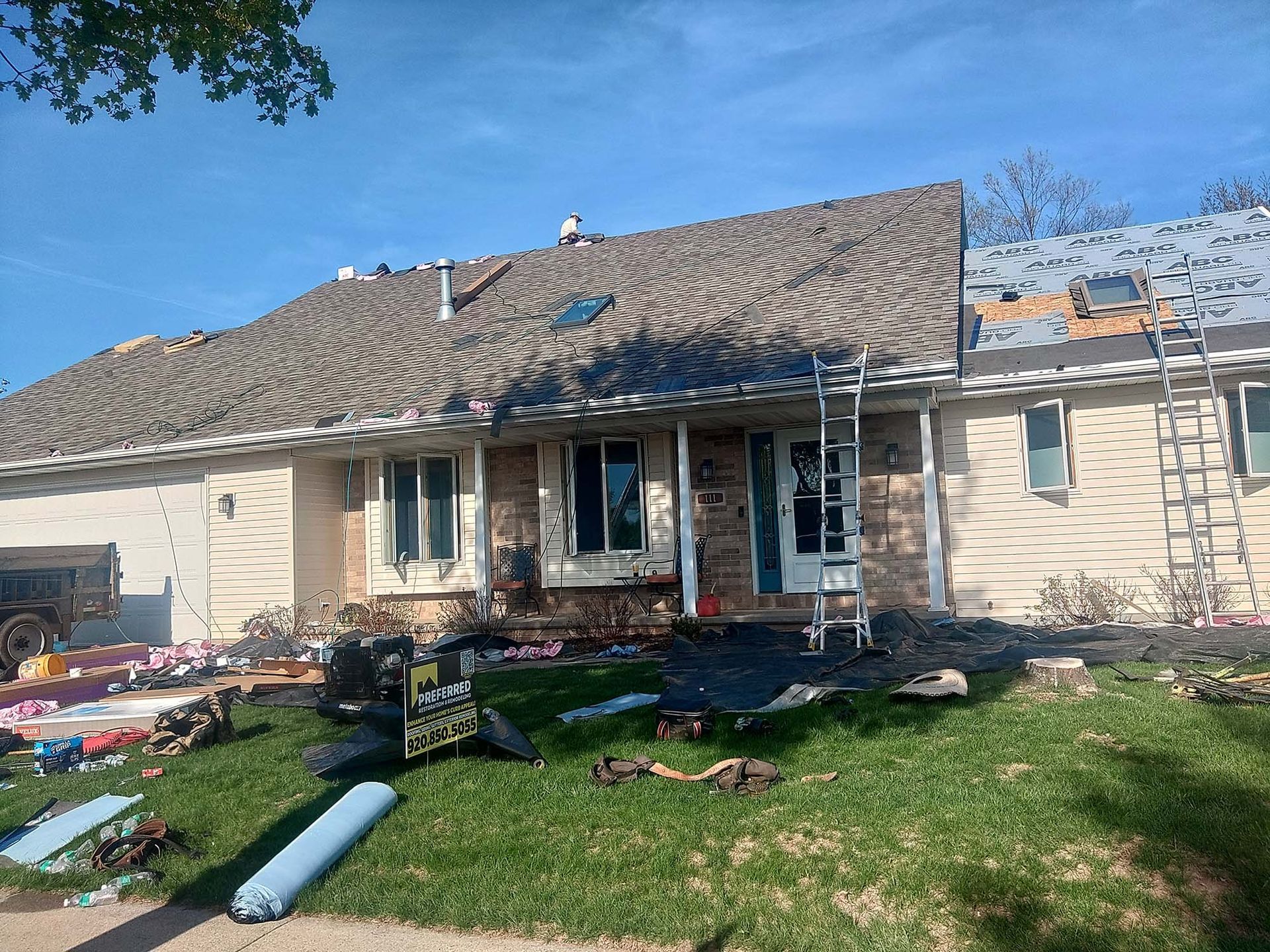 House with roof partially replaced, ladder leaning against it. Debris on lawn, clear sky.