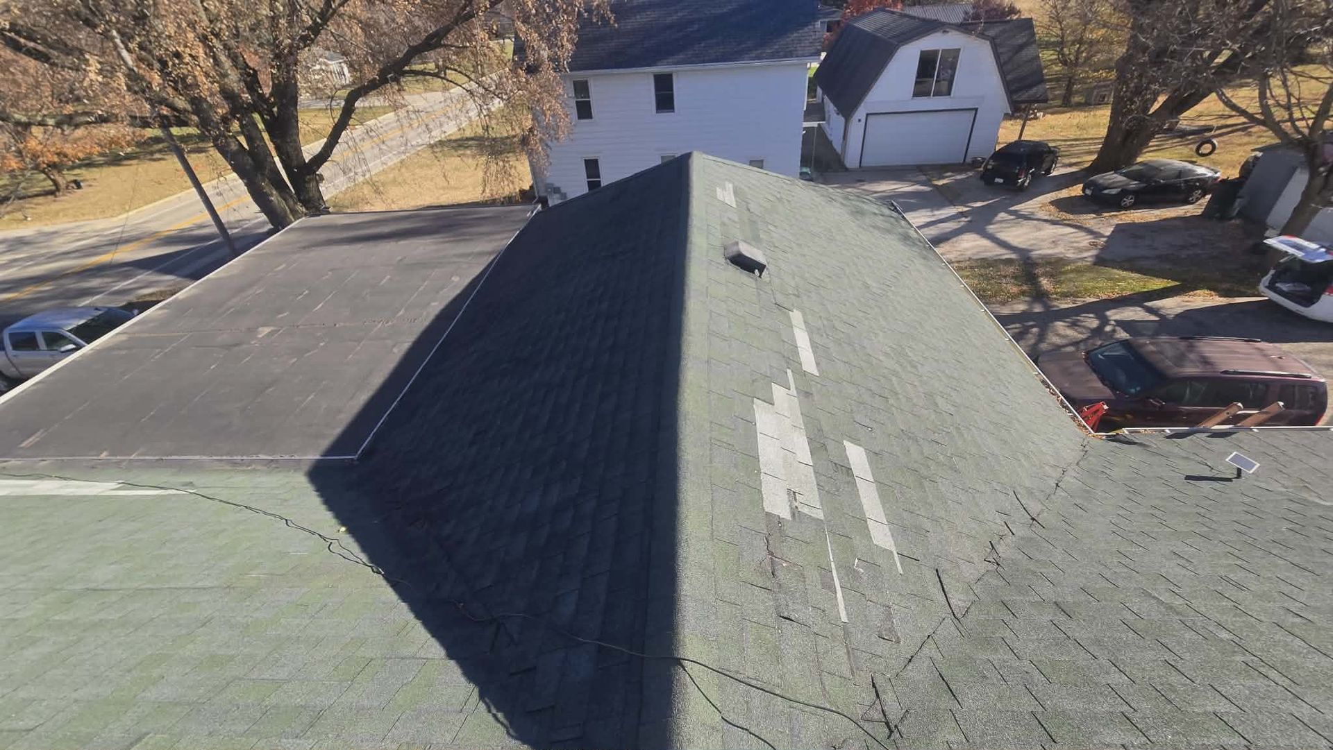 View of a green, weathered roof with patches, trees, and buildings in the background on a sunny day.