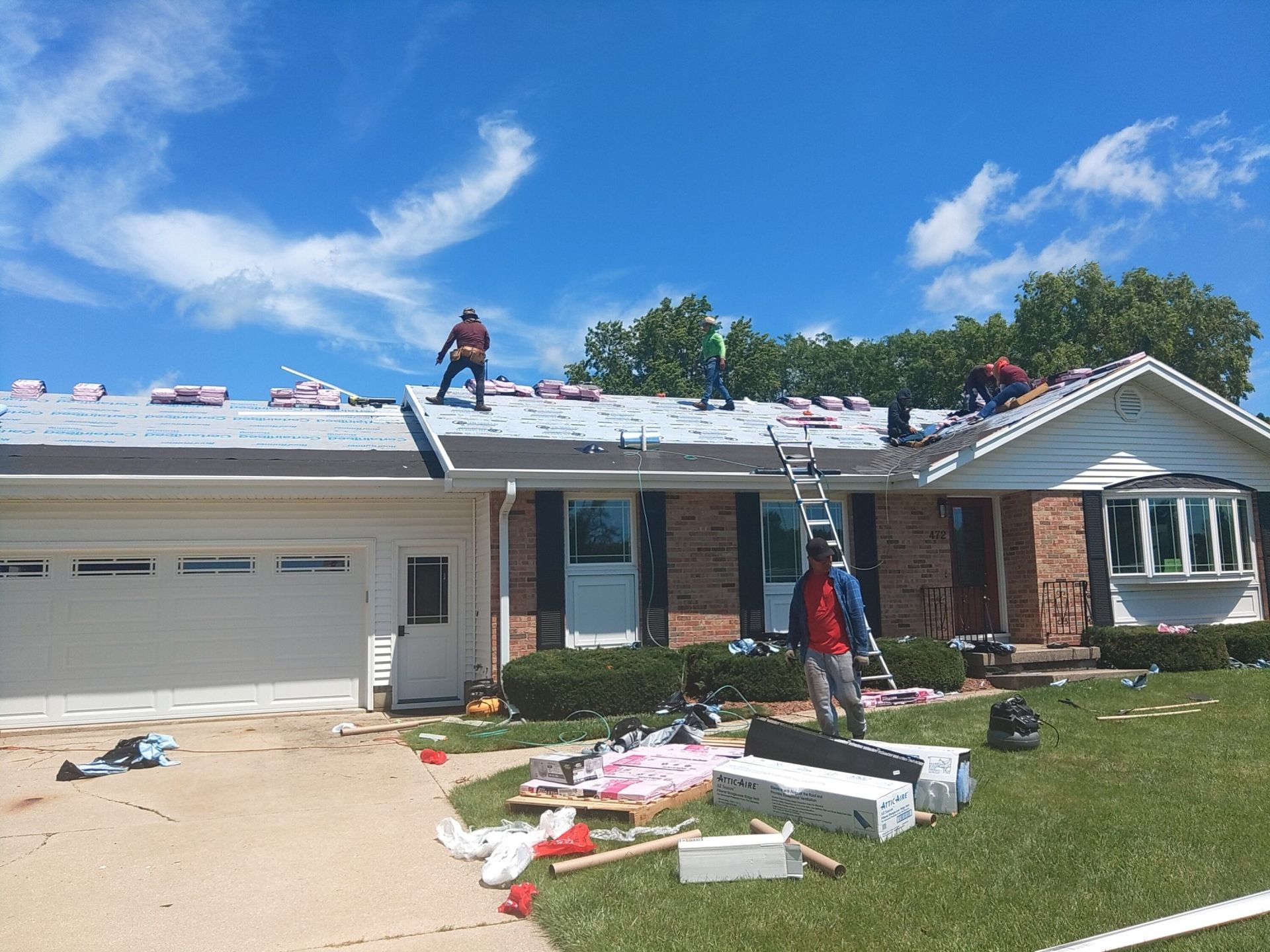 Roofers working on a residential roof under a blue sky. Materials and a ladder are on the ground.