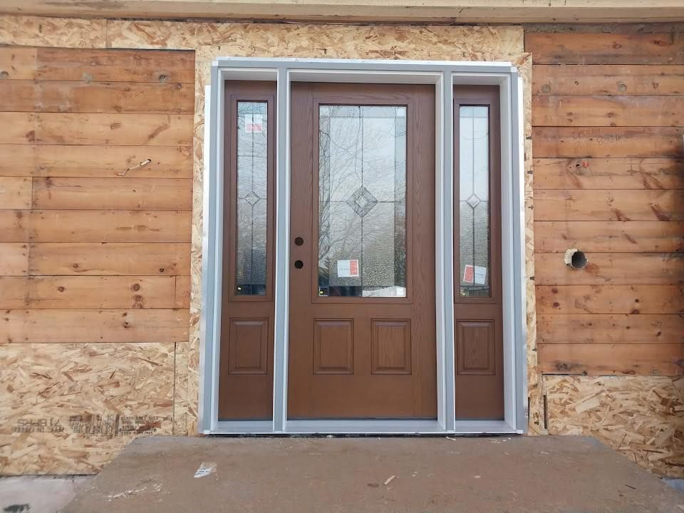 Brown front door with sidelights, set in unfinished wooden siding.