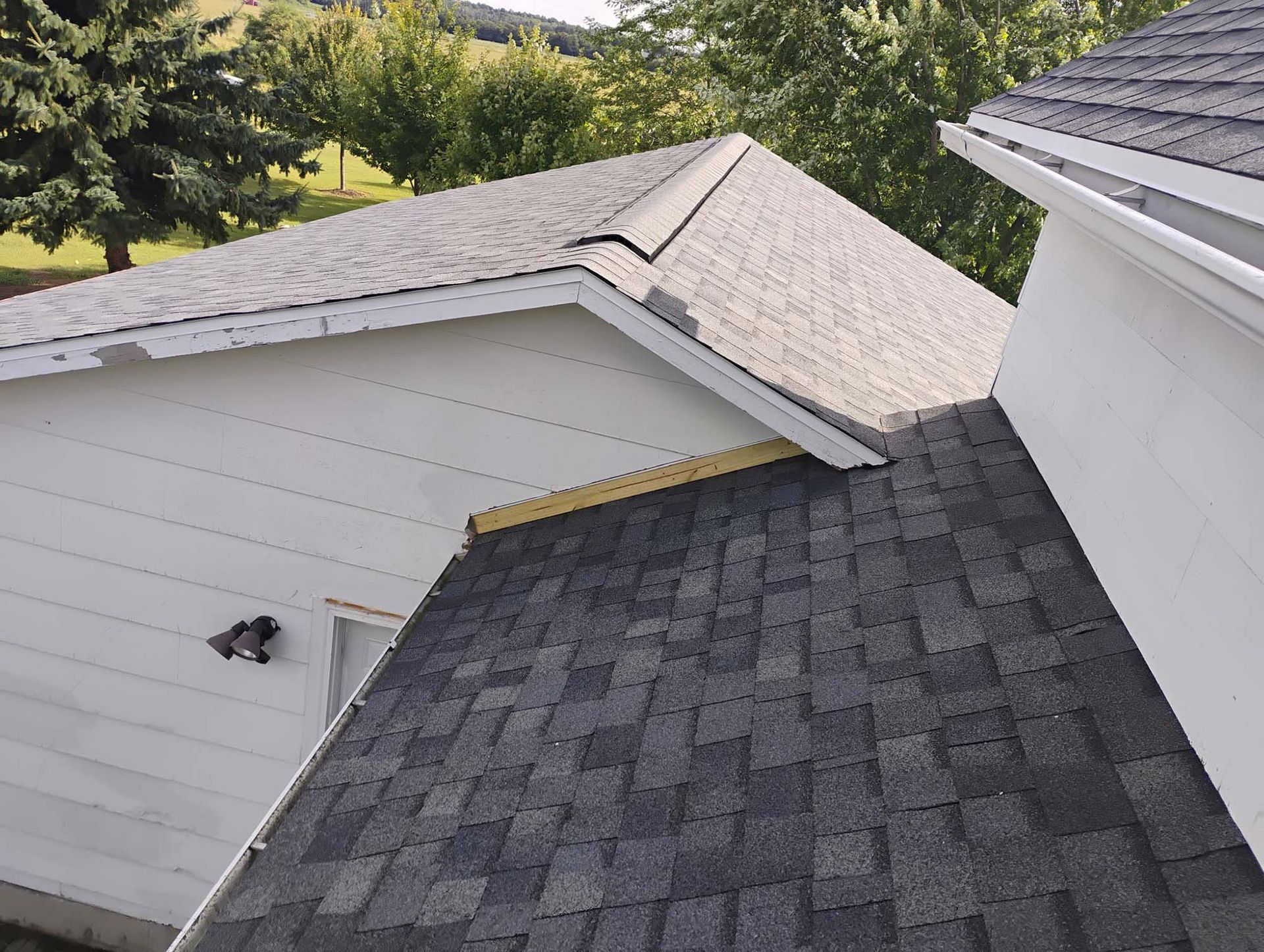 Roof of a white house with grey shingles. Damaged roof area and trees are visible in the background.