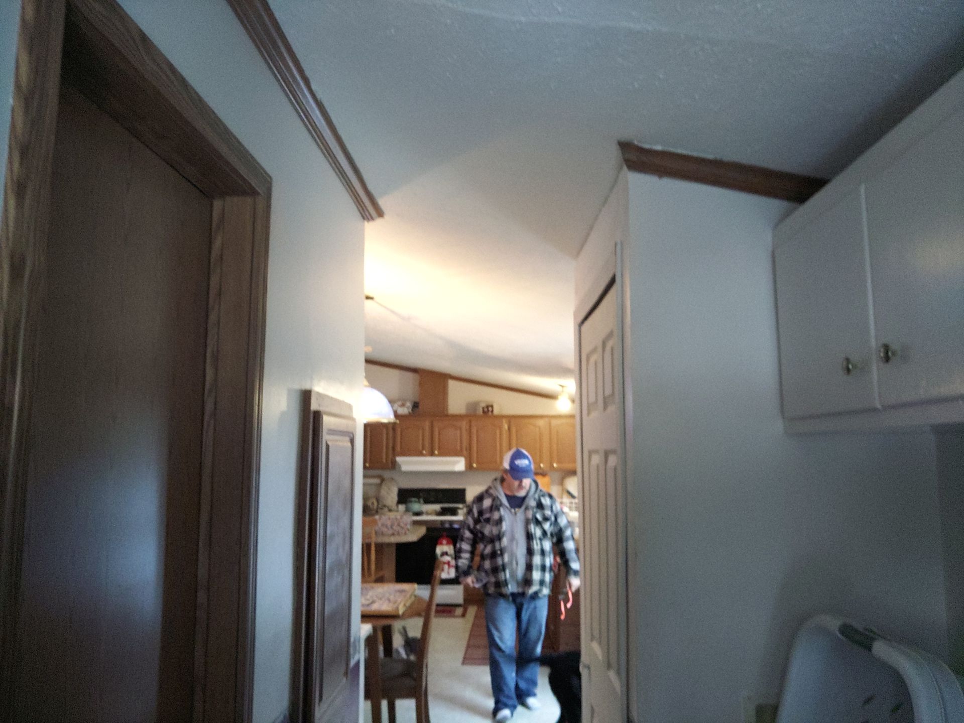 Man walking through doorway into kitchen. He wears a plaid shirt and hat. Kitchen cabinets and table visible.