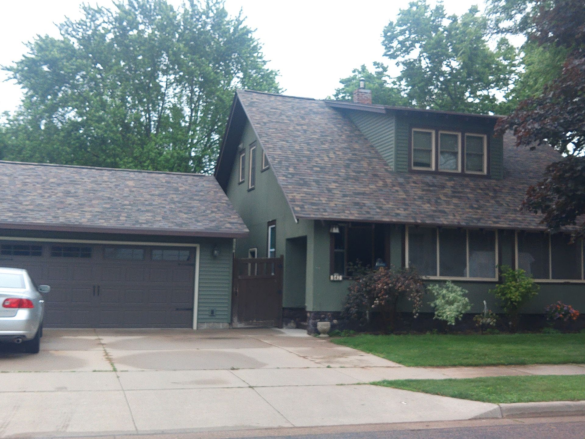 Green house with attached garage, car in driveway, surrounded by trees.