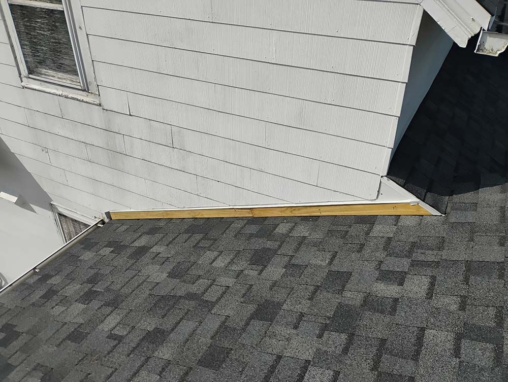 Overlapping roof shingles meet a white house wall. Wooden trim lines the roof edge.