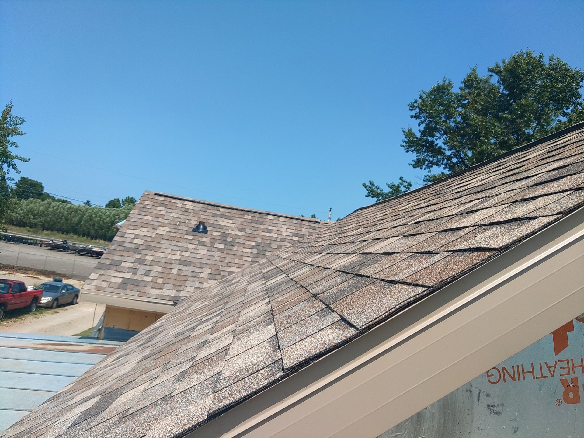 Roof with brown shingles, blue sky, and a glimpse of cars and trees in the background.