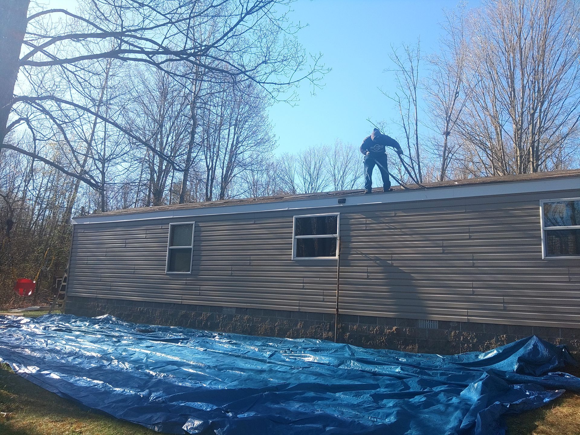 Person on a mobile home roof. Blue tarp on the ground. Trees in the background, blue sky.