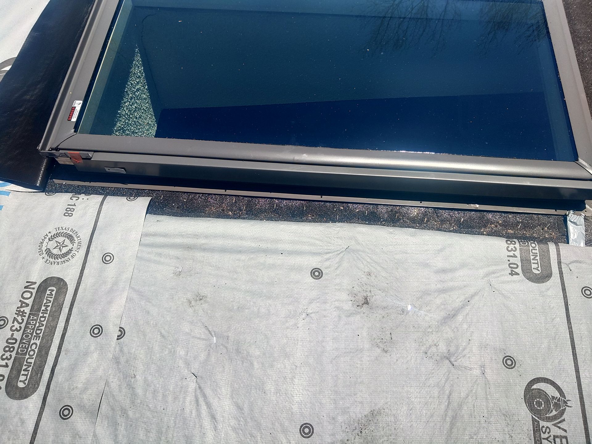 Skylight installed on a roof with roofing underlayment visible. Brown frame, dark glass reflecting the sky.