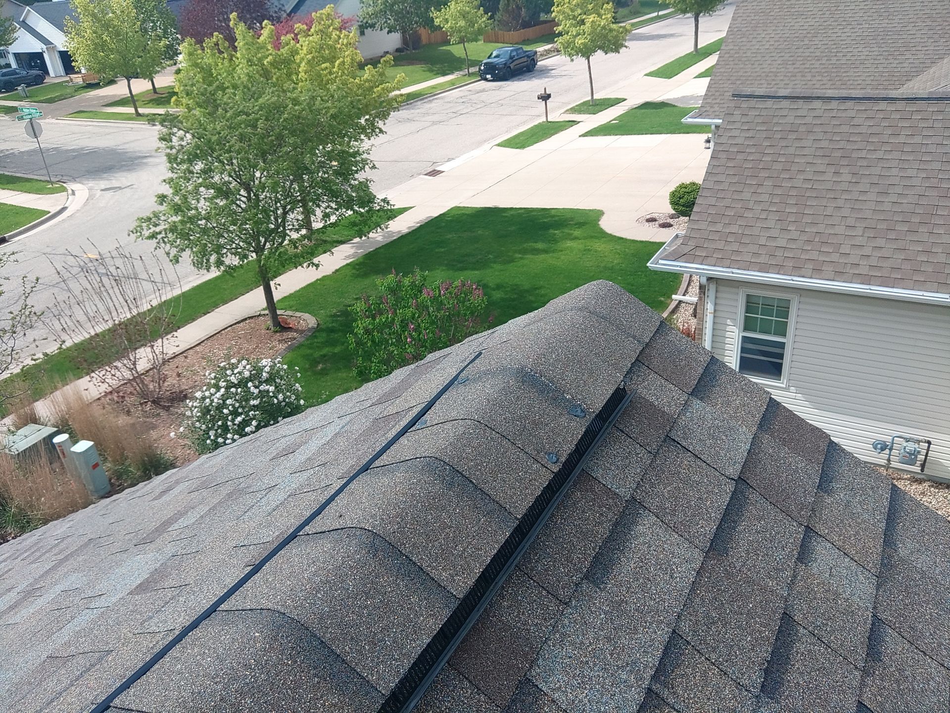 Rooftop view of asphalt shingles, overlooking a suburban neighborhood with green lawns and trees.
