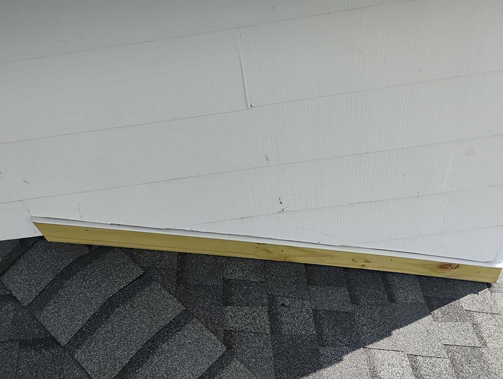 A wooden board rests on a dark gray shingled roof, below white siding.