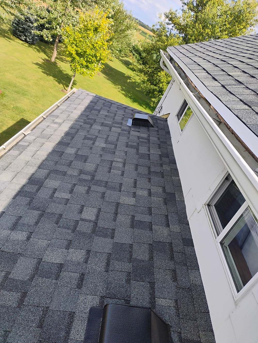 A roof with gray shingles, a vent, and a white house, seen from above. Green trees in the background.