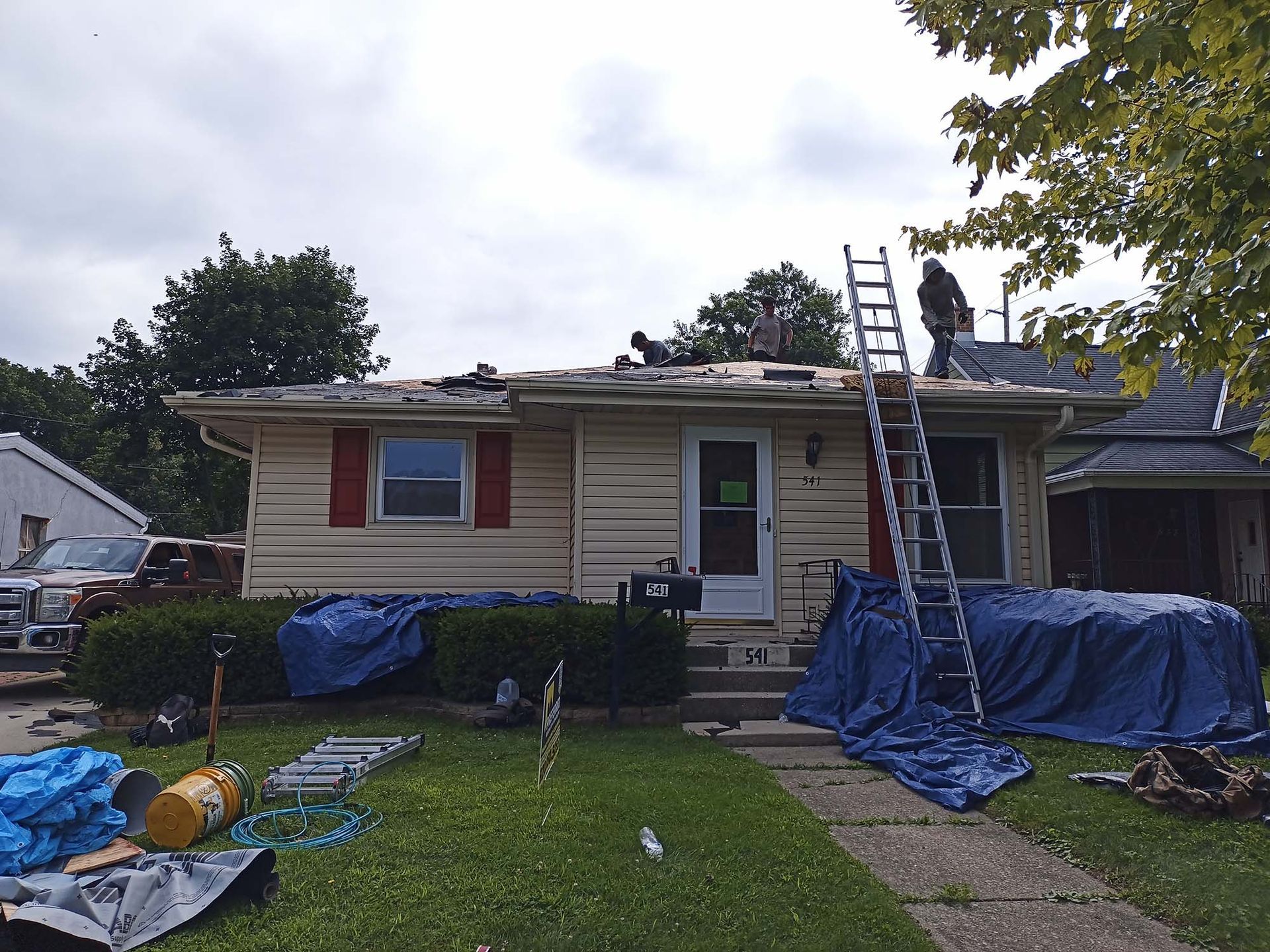 House with fire damage; workers on roof with ladder; blue tarps cover items.