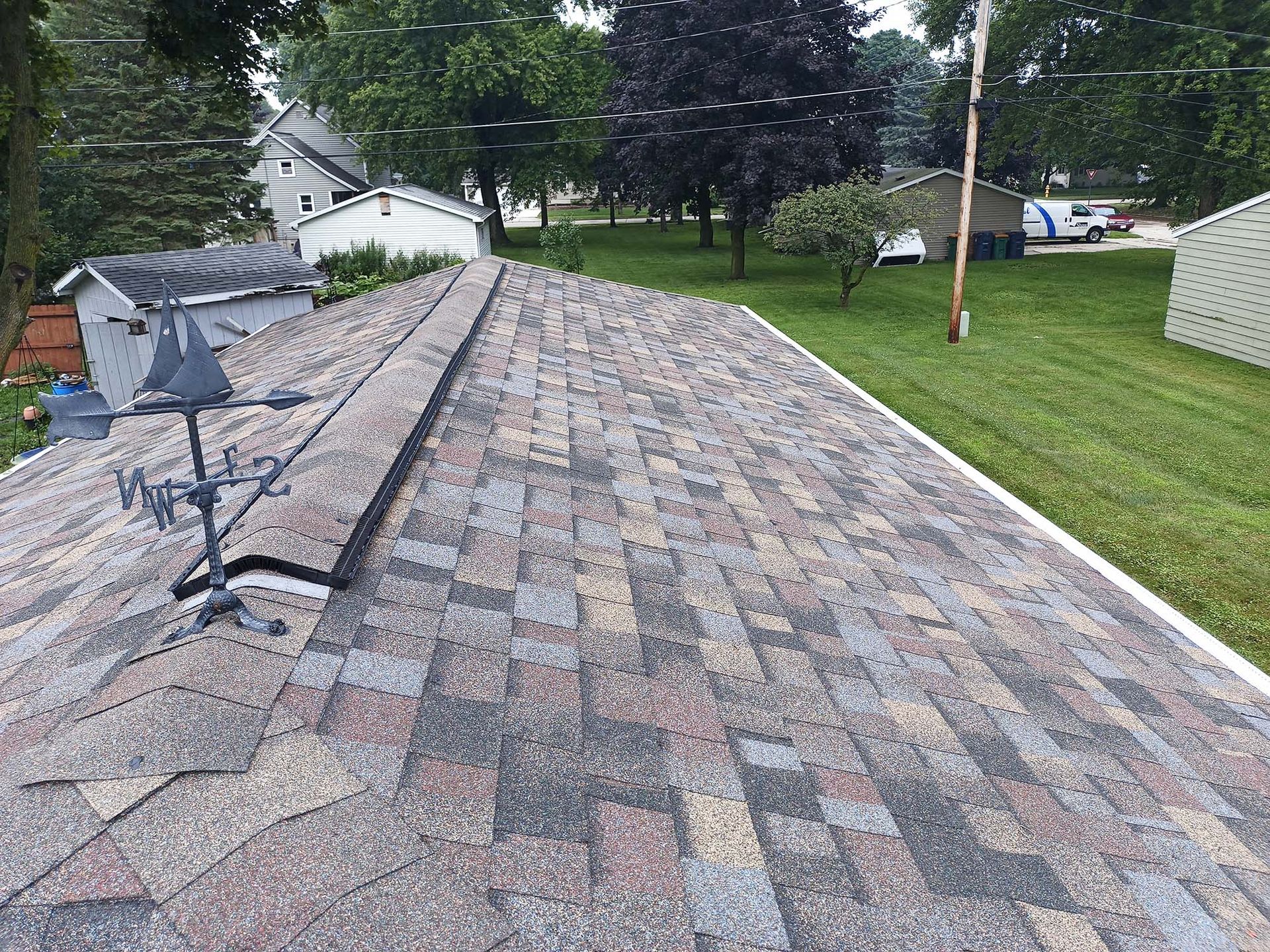 A multi-colored shingled roof with a satellite dish and neighboring houses in a green yard.