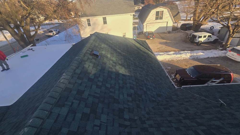 Rooftop view of a dark green shingled roof with a hole. White snow on a neighboring roof and yard.