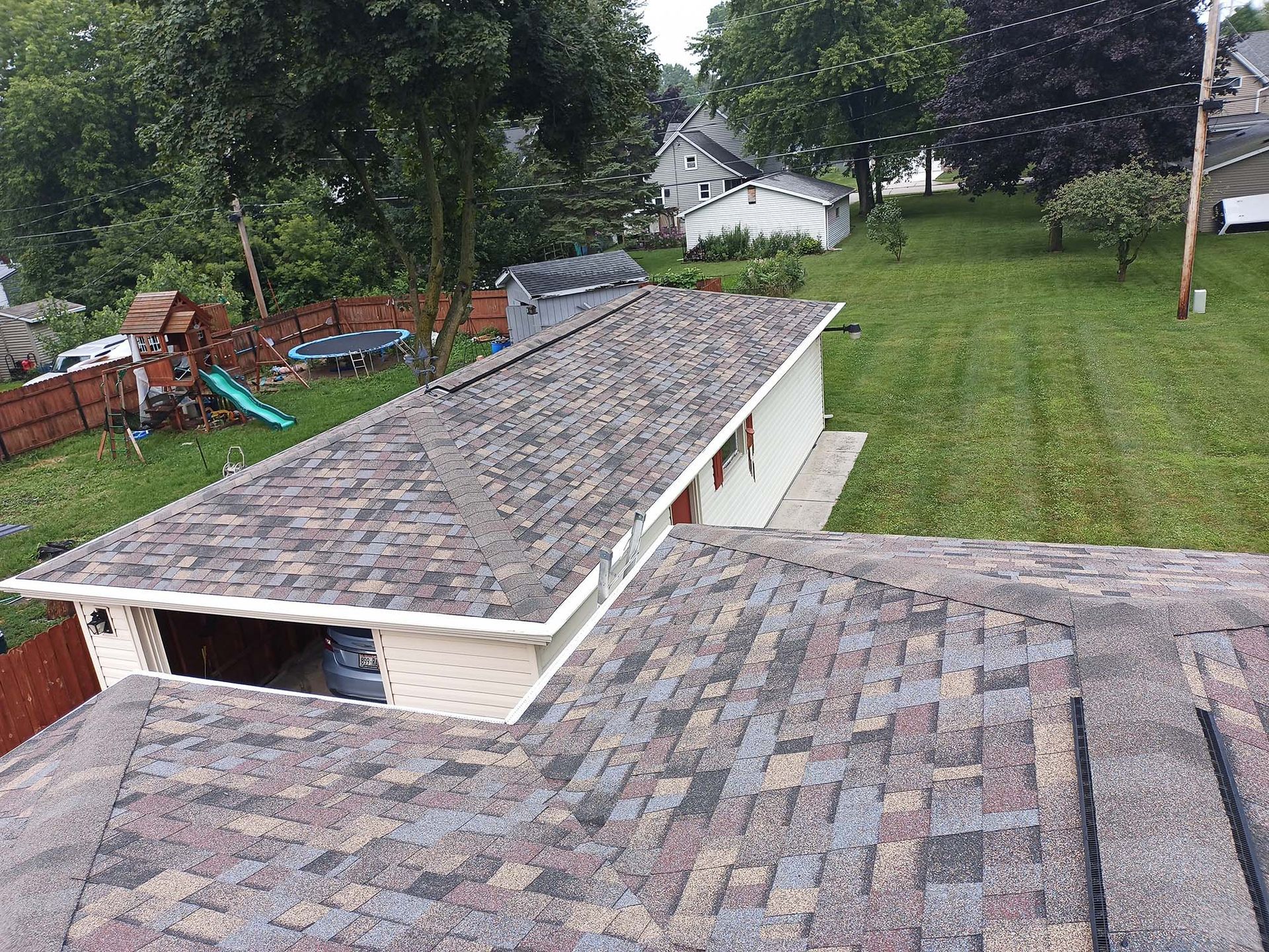 Overhead view of a house roof with a garage in the backyard, green grass, and trees.