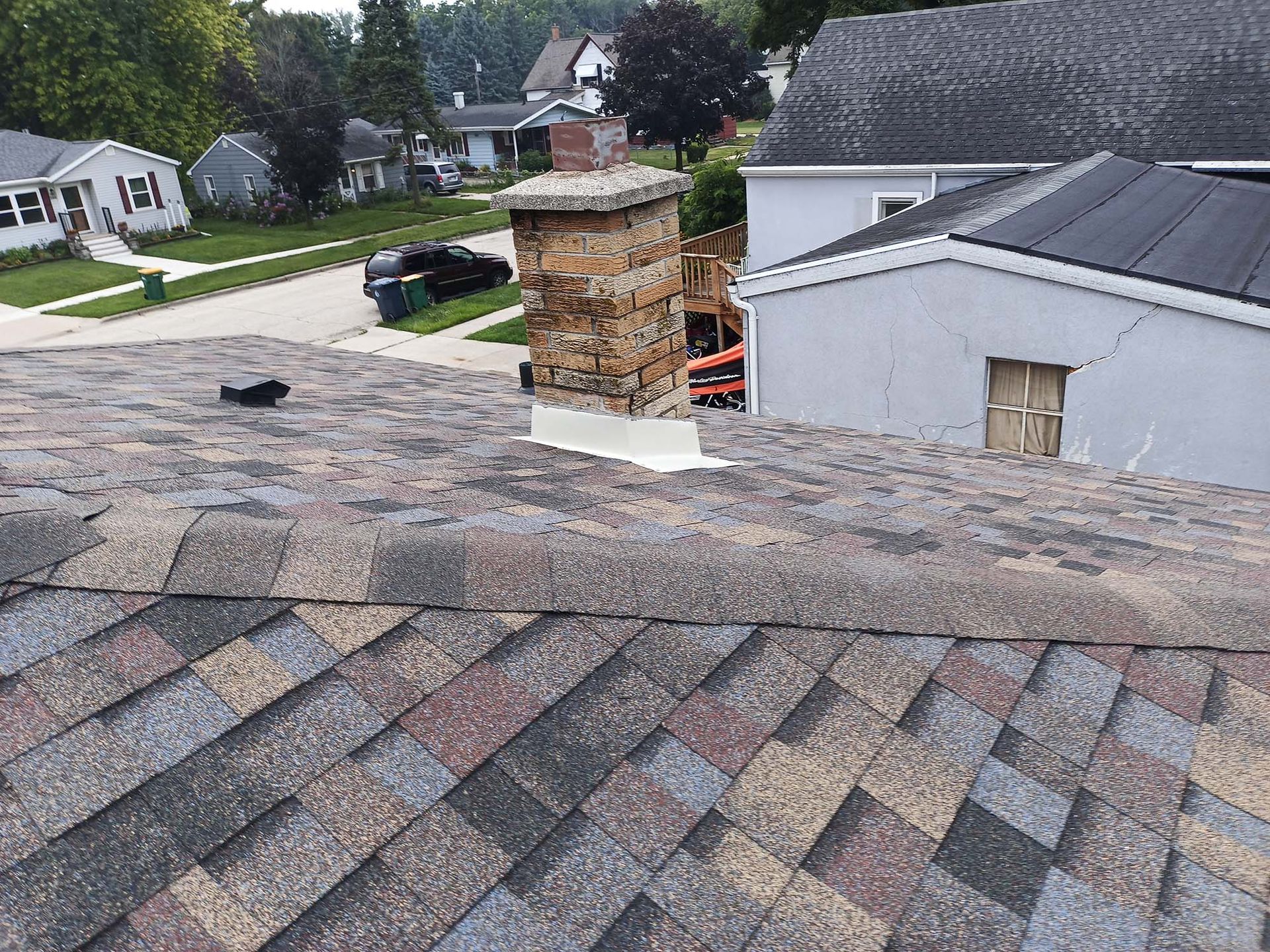 Rooftop view with a damaged brick chimney, surrounded by asphalt shingles. Houses and street in background.