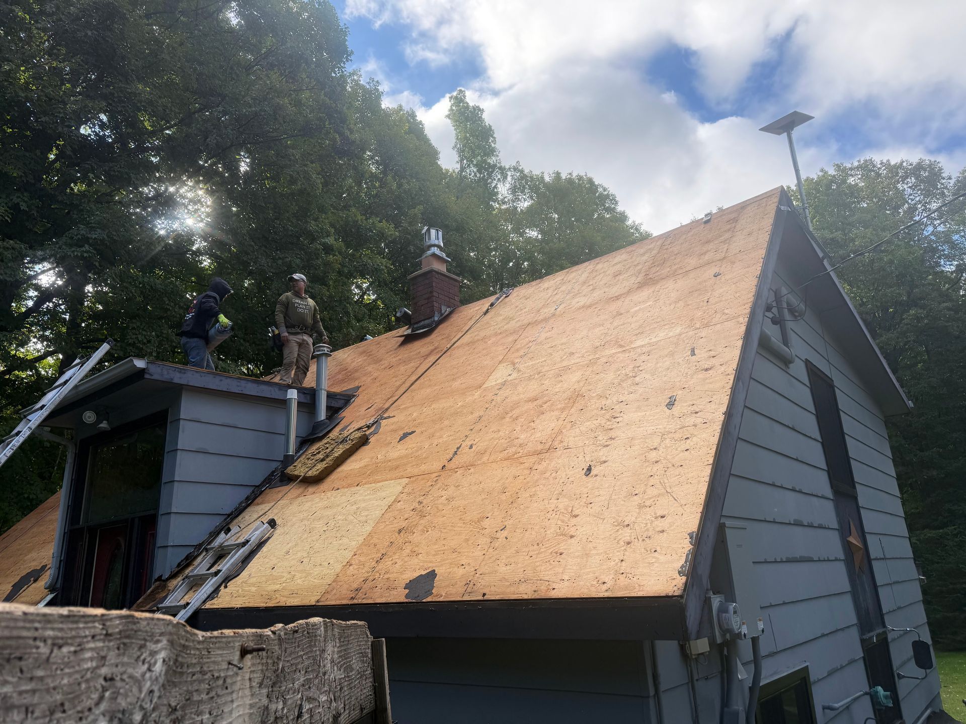 Roofers working on a house with exposed plywood, a chimney, and a cloudy sky overhead.