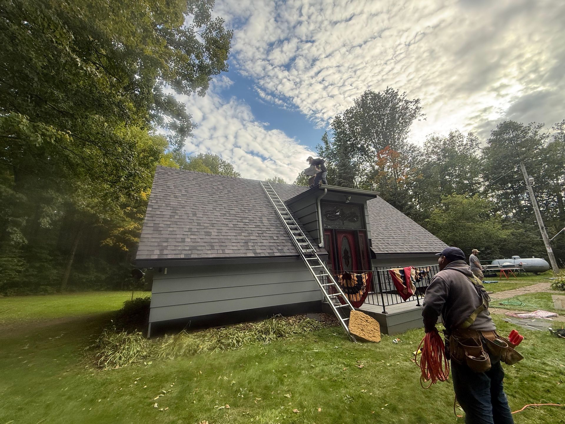 Workers on roof, one on ladder, with tools. Cloudy sky and grassy yard.