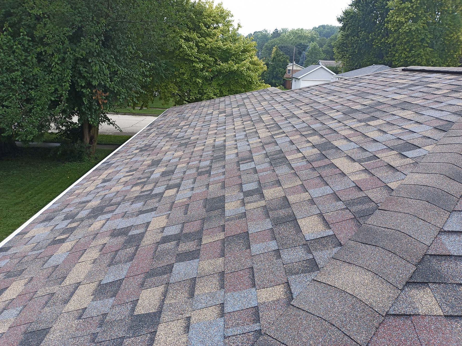 Close-up view of a multi-colored asphalt shingle roof. Trees and grass in the background.