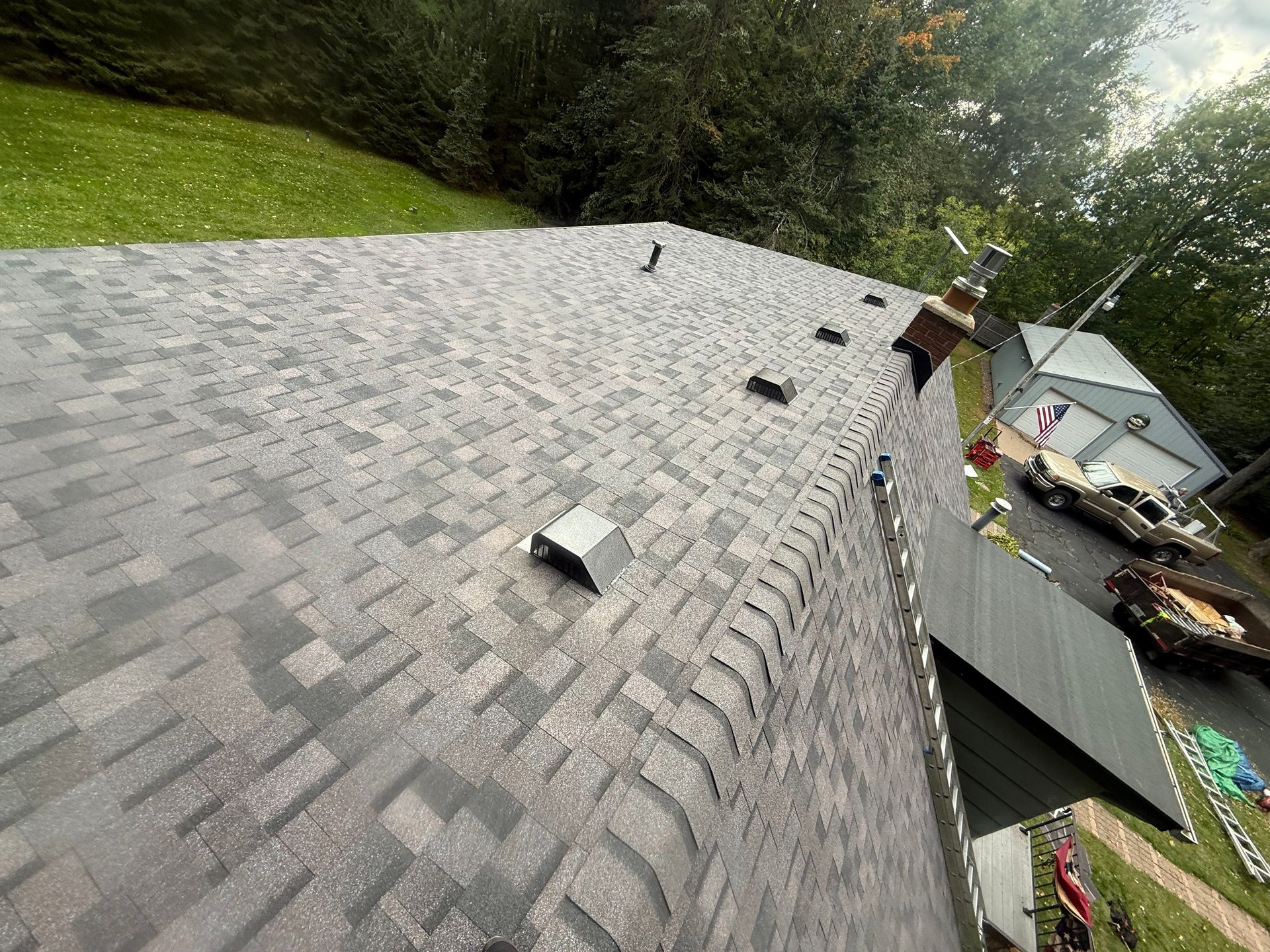 Overhead view of a gray shingled roof with several vents. A detached garage and vehicles are in the background.
