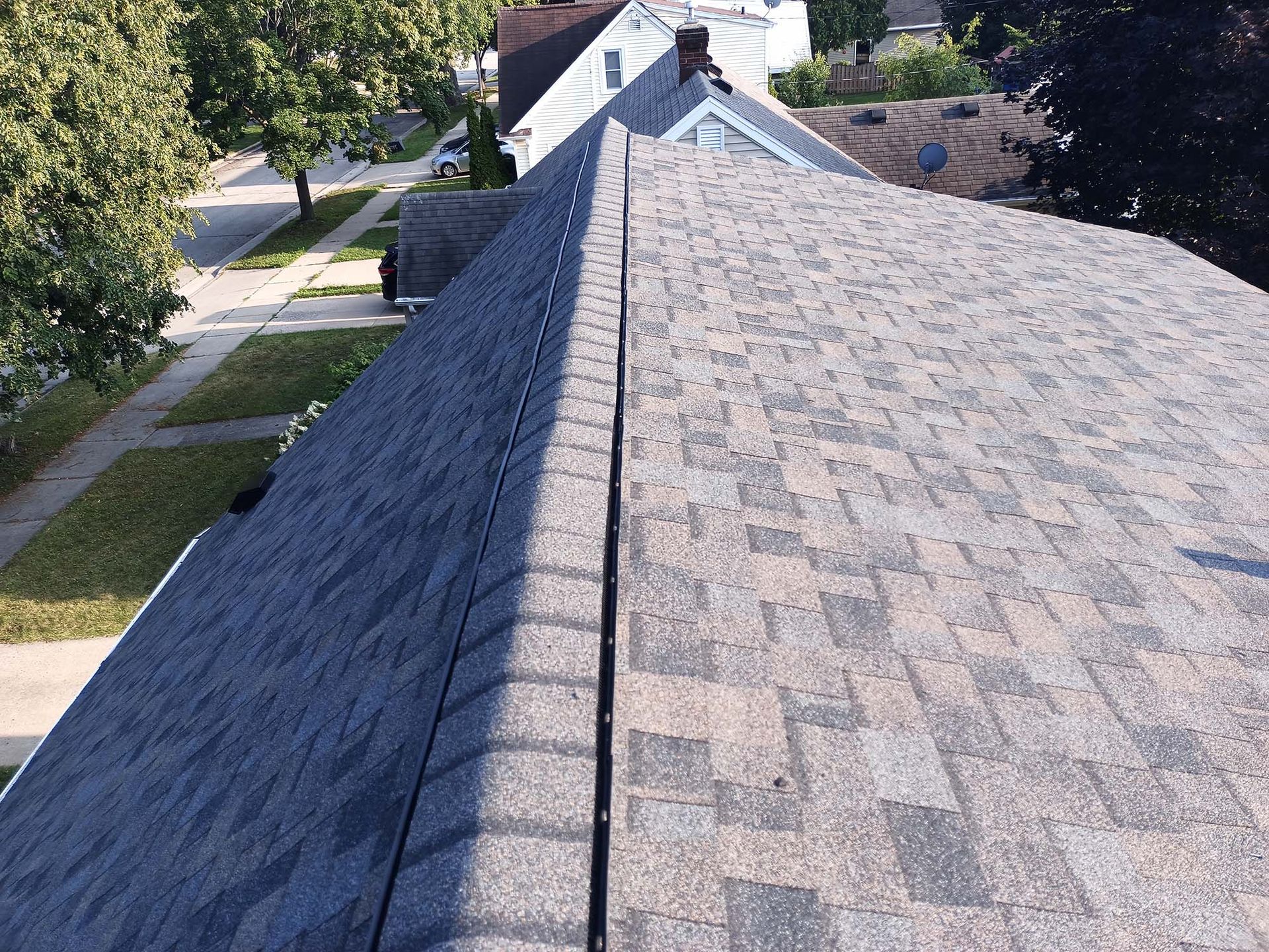 Overhead view of a roof with grey asphalt shingles, a visible ridge, and surrounding houses.