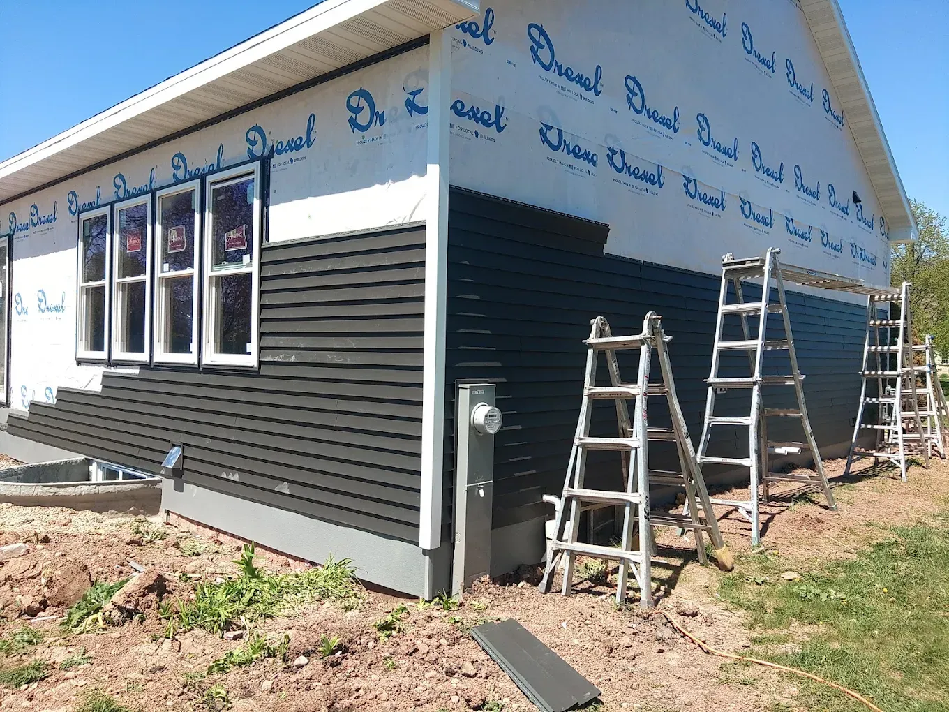 A house under construction; dark siding is being installed. Several ladders are leaning against the house.
