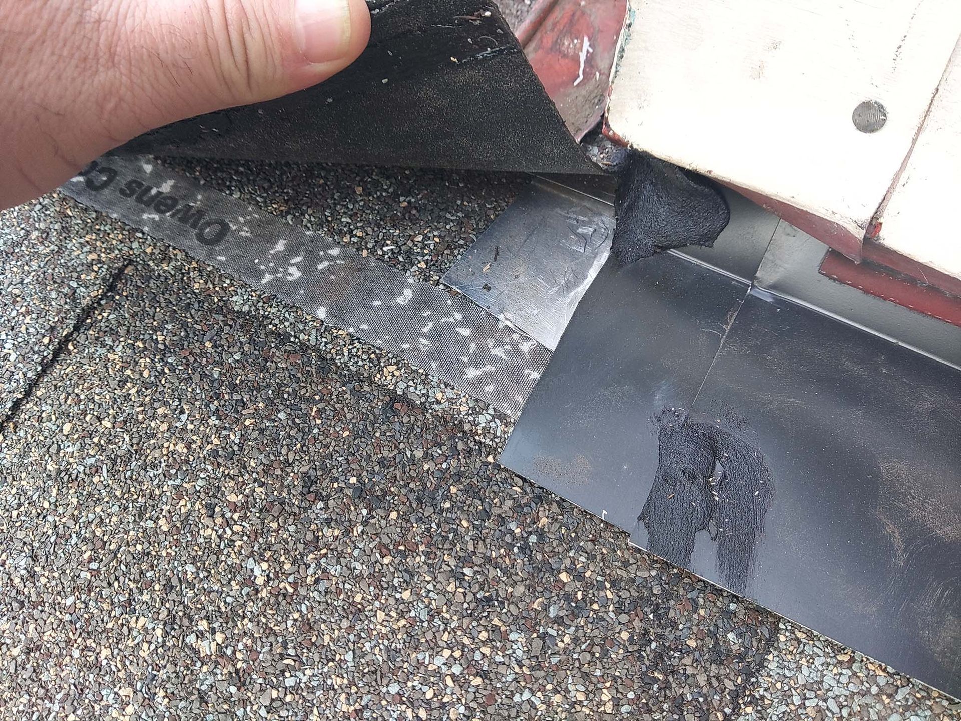 A person's hand lifts roofing material to expose metal flashing next to a wall.