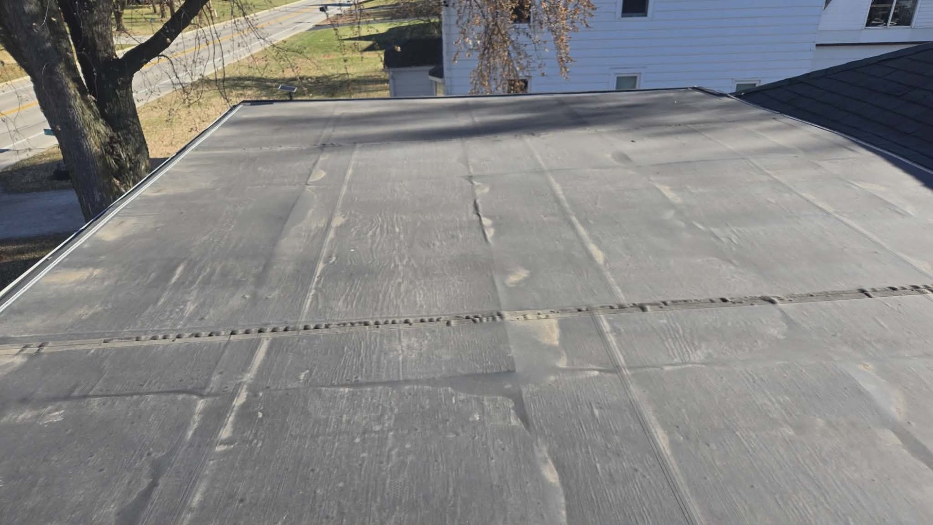 Flat, dark-colored roof with visible seams. Building and tree visible in the background on a sunny day.