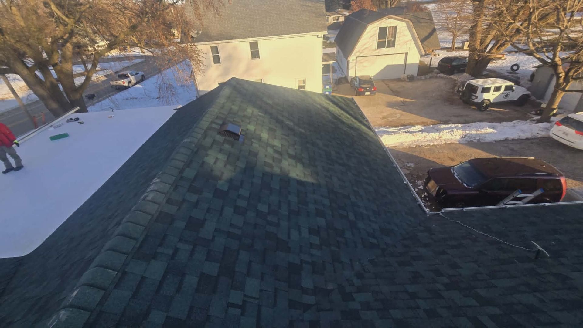 A roof with dark shingles is being worked on, with snow on another roof and vehicles in the background.