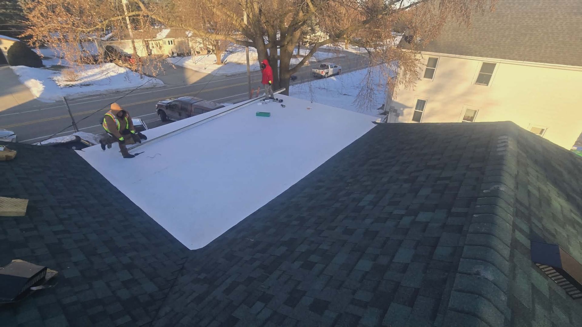 Person clearing snow from a roof. The roof has green shingles, and a snow-covered section with trees and buildings in the background.