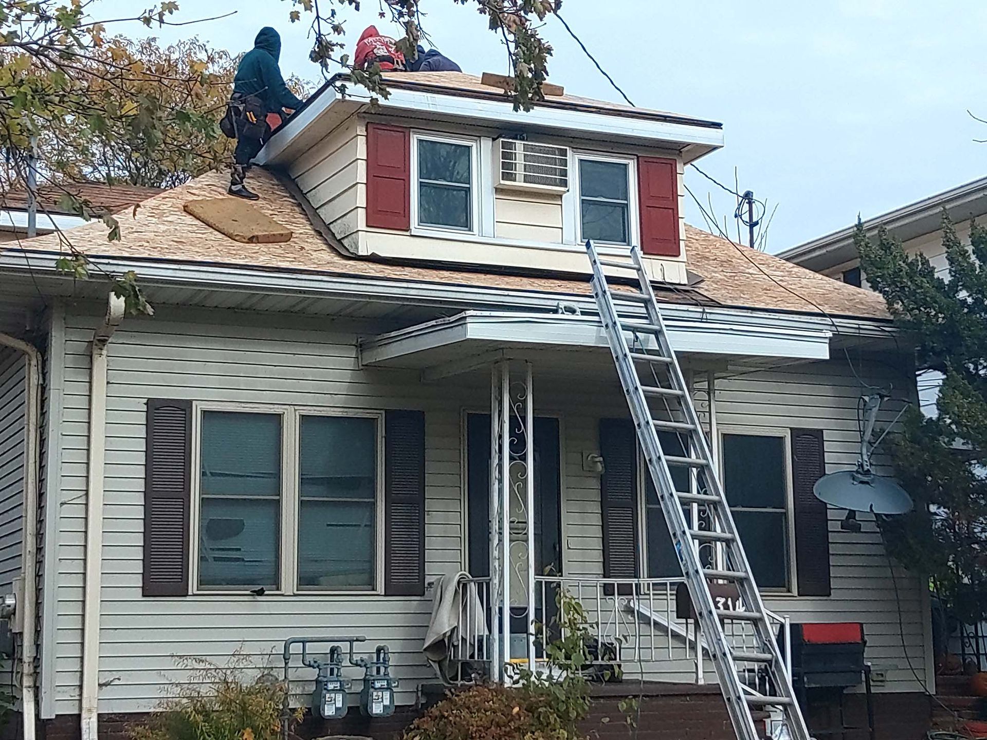 Workers on a house roof replacing shingles, ladder propped on porch.